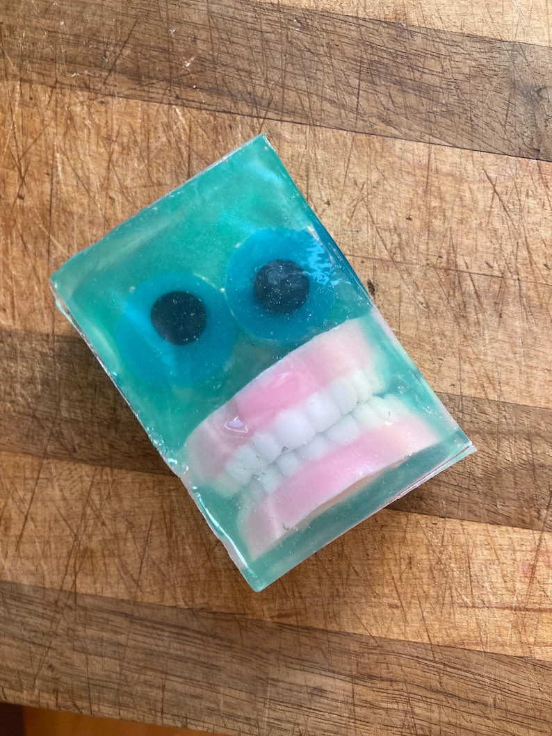 May include: A rectangular bar of soap with a teal translucent base. Two blue, round candies with black centers are set near the top. A set of pink-based dentures is embedded in the soap.