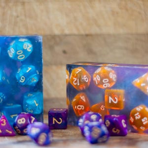 May include: Two resin dice sets, one blue and one purple and orange, with white numbering. The dice are encased in rectangular blocks. Additional loose dice in purple and blue are scattered in front of the blocks. The background is a wooden surface.