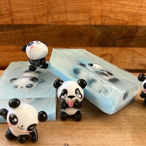 May include: Four small panda figurines, two are on top of clear blue soap bars, one is on a white soap bar, and one is standing on a wooden surface. The soap bars have panda figurines embedded in them.