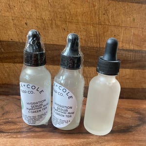 May include: Three clear glass bottles with black dropper tops and white labels. The labels say "K+COLE SOAP CO." and "HYDRATION SERUM Hyaluronic Acid & GREEN TEA".
