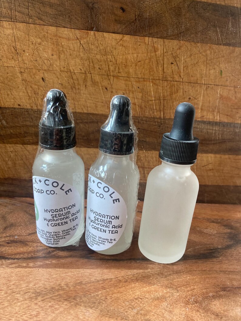 May include: Three clear glass bottles with black dropper tops and white labels. The labels say "K+COLE SOAP CO." and "HYDRATION SERUM Hyaluronic Acid & GREEN TEA".