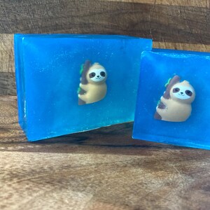 May include: Two blue soap bars with a sloth design. The sloth is brown and has a green leaf in its hand.