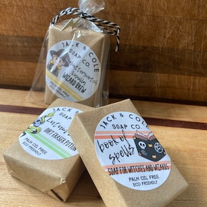 May include: Three bars of handmade soap with Halloween-themed labels. The first bar is labeled "Butterscotch Vanilla Wizard Brew", the second bar is labeled "Lutum Dirt Eraser Potion", and the third bar is labeled "Book of Spells Soap for Witches and Wizards". All three bars are palm oil free and eco-friendly.