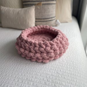 May include: A circular pet bed in a soft pink hue, crafted from thick, knitted yarn. The bed features a raised rim and is placed on a white bedspread. Pillows are visible in the background.