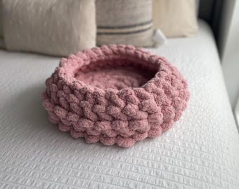 May include: A circular pet bed in a soft pink hue, crafted from thick, knitted yarn. The bed features a raised rim and is placed on a white bedspread. Pillows are visible in the background.