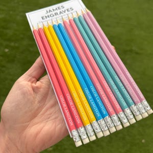 Op de afbeelding: Een set van acht houten potloden gerangschikt op een witte kaart met het label JAMES ENGRAVES. De potloden hebben een kleurverloop van roze, geel, blauw en turquoise, elk gepersonaliseerd met de naam CHLOE LANGTON in witte tekst en voorzien van een zilveren huls en een witte gum.