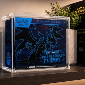 May include: A clear acrylic display case containing a blue Pokémon Elite Trainer Box. The box features a dark blue design with a light blue outline of a Charizard, the text Mega Evolution Phantasmal Flames, and a warning label at the bottom.