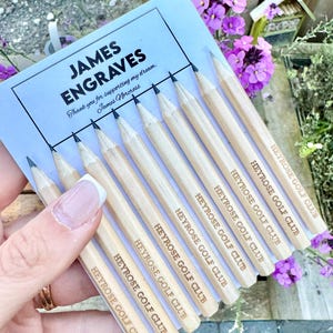 Puede incluir: Un conjunto de diez lápices de madera con el texto "HEYROSE GOLF CLUB" grabado en cada lápiz. Los lápices están dispuestos en forma de abanico y se sostienen en una mano.