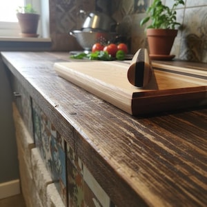 May include: A wooden chopping board with a dark handle and a light wood surface rests on a rustic wooden countertop. The countertop has a distressed finish. A small terracotta pot with a green plant is in the background.