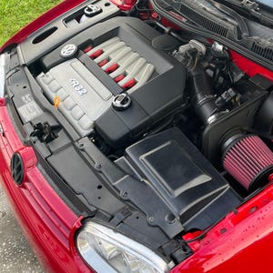 May include: Close-up of a red Volkswagen Golf GTI engine bay with a black air intake and a red air filter. The engine is a turbocharged 1.8T engine with a silver and black cover. The engine cover has the text "VR6" on it.