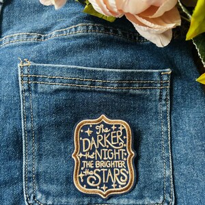 May include: A rectangular embroidered patch with a dark blue background and gold border, featuring the text "The DARKER the NIGHT the BRIGHTER the STARS" in gold lettering, attached to the back pocket of a pair of blue denim jeans.