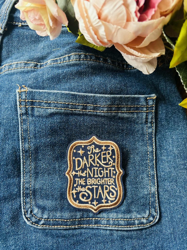 May include: A rectangular embroidered patch with a dark blue background and gold border, featuring the text "The DARKER the NIGHT the BRIGHTER the STARS" in gold lettering, attached to the back pocket of a pair of blue denim jeans.