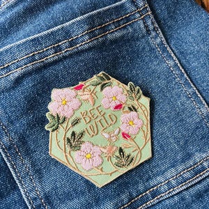 Puede incluir: Un parche hexagonal con el texto "BEE WILD" rodeado de flores rosas, hojas verdes y una peque&ntilde;a abeja. El parche est&aacute; sobre un fondo verde claro y cosido en un bolsillo de mezclilla azul.