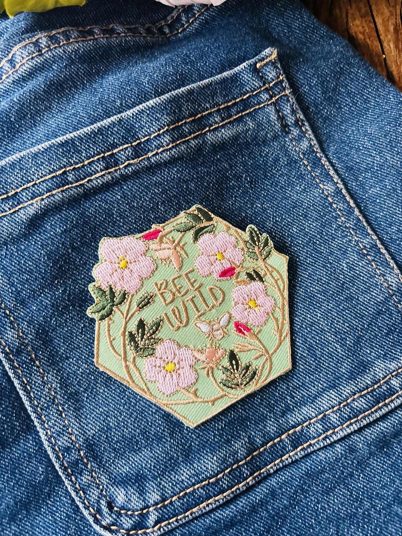 Puede incluir: Un parche hexagonal con el texto "BEE WILD" rodeado de flores rosas, hojas verdes y una peque&ntilde;a abeja. El parche est&aacute; sobre un fondo verde claro y cosido en un bolsillo de mezclilla azul.
