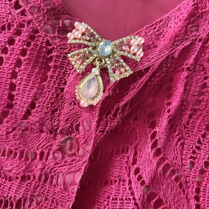 Puede incluir: Una tela de encaje rosa con un broche de lazo dorado y rosa adornado con joyas. El broche tiene un colgante en forma de lágrima con una piedra rosa.