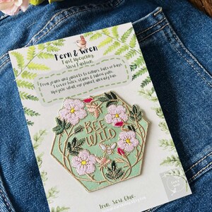 Puede incluir: Un parche hexagonal con la frase "BEE WILD" rodeada de flores rosas y hojas verdes. El parche est&aacute; sobre una tarjeta con el texto "Fern & Wren" e informaci&oacute;n sobre el reciclaje, sobre un fondo de mezclilla.