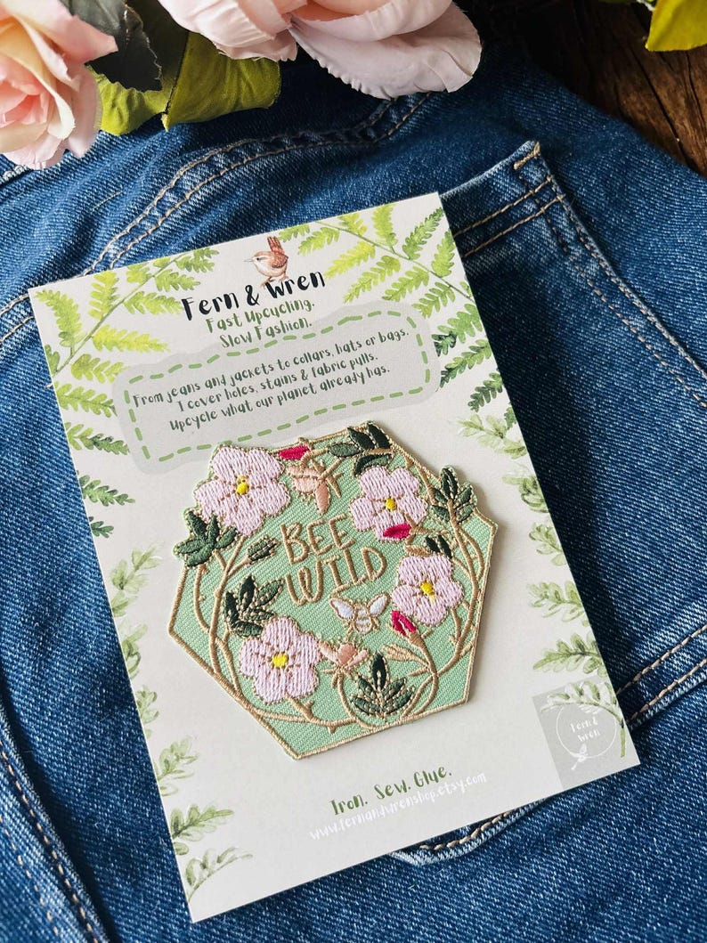 Puede incluir: Un parche hexagonal con la frase "BEE WILD" rodeada de flores rosas y hojas verdes. El parche est&aacute; sobre una tarjeta con el texto "Fern & Wren" e informaci&oacute;n sobre el reciclaje, sobre un fondo de mezclilla.