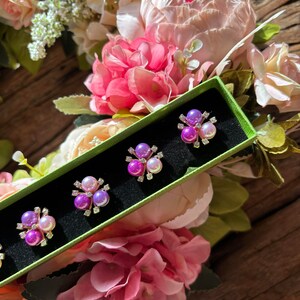 May include: A green box with four purple and pink pearl flower brooches. The box is surrounded by pink and white flowers.