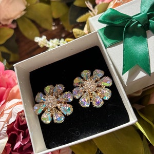 May include: A pair of iridescent flower-shaped earrings with clear rhinestones in a white gift box with a green bow.