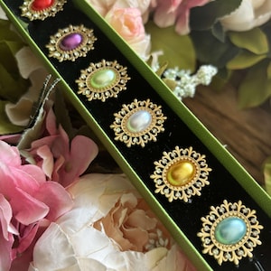 May include: A set of six gold-toned napkin rings with oval-shaped glass stones in various colors, including red, purple, green, white, yellow, and blue. The rings are displayed in a green box with a black interior.