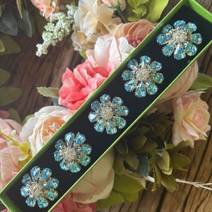 Puede incluir: Un juego de seis anillos de servilleta en forma de flor azul y plata en una caja verde. Los anillos están hechos de diamantes de imitación y están dispuestos en una fila.