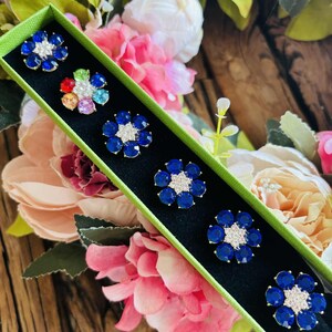 May include: A green box displays six flower-shaped brooches. Five brooches have blue petals and a white center. One brooch has multicolored petals. The brooches are set against a black background, surrounded by pink and white flowers.