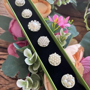 May include: A set of six pearl and rhinestone brooches in a green box. The brooches are arranged in a row, with the largest brooch at the center. The brooches are all round and have a silver finish.