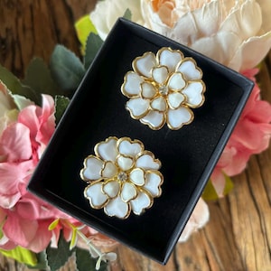 May include: A pair of white and gold flower-shaped earrings displayed in a black box. The earrings have a gold outline and a small crystal in the center. The background features pink and white flowers on a wooden surface.
