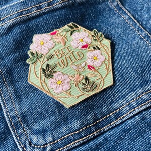 Puede incluir: Un parche hexagonal con las palabras "BEE WILD" bordadas sobre un fondo verde claro. El parche presenta flores rosas, hojas verdes y un borde dorado, sobre un fondo de mezclilla azul.