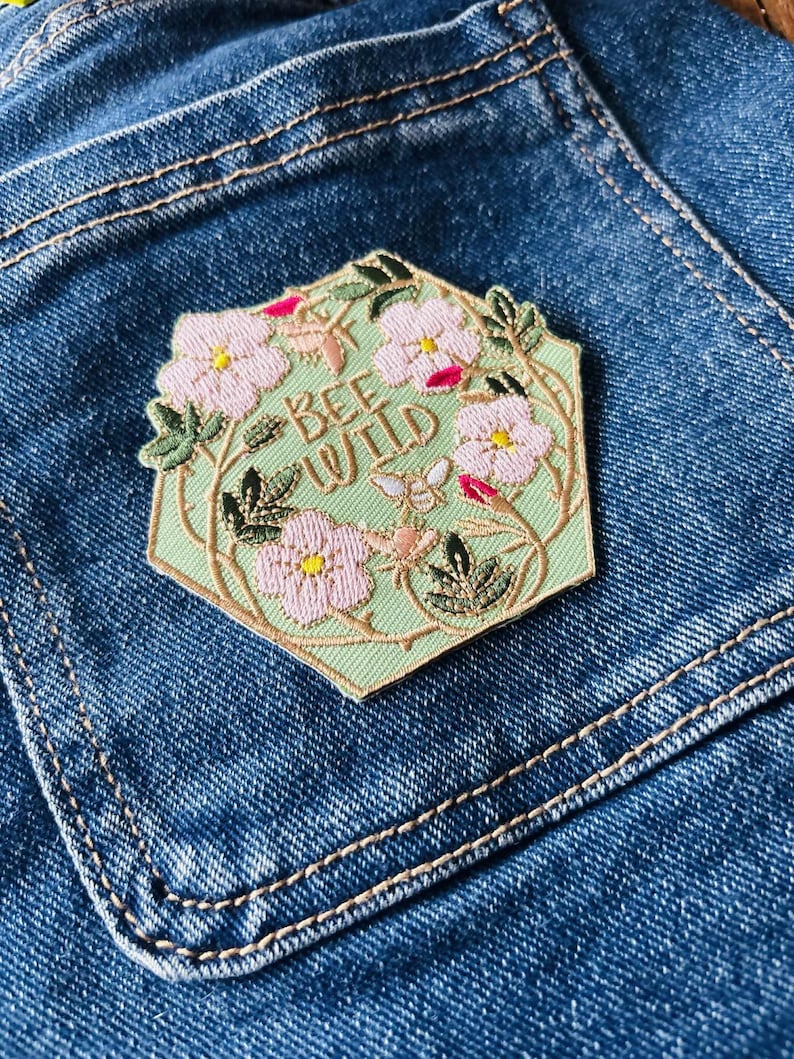 Puede incluir: Un parche hexagonal con las palabras "BEE WILD" bordadas sobre un fondo verde claro. El parche presenta flores rosas, hojas verdes y un borde dorado, sobre un fondo de mezclilla azul.