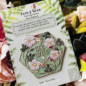 Puede incluir: Un parche bordado con un dise&ntilde;o floral y las palabras "BEE WILD". El parche presenta flores rosas, hojas verdes y una abeja sobre un fondo verde claro. El parche est&aacute; en una tarjeta con el texto "Fern & Wren, Fast Upcycling, Slow Fashion."