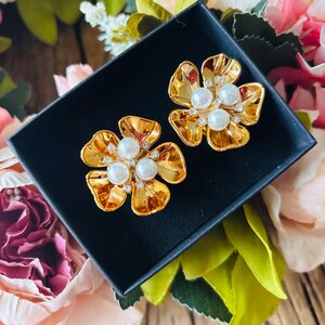 May include: A pair of gold-toned flower-shaped earrings with pearl and crystal accents, presented in a black box. The earrings have a detailed petal design and are set against a backdrop of pink and green floral arrangements.