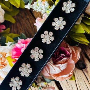May include: Six flower-shaped brooches with clear crystals and gold-colored metal settings are displayed in a black box. The brooches are arranged in a row. The background features a wooden surface and colorful flowers.