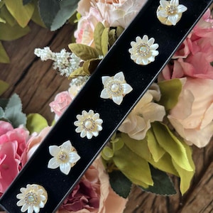 May include: A set of seven flower-shaped brooches in a black box. The brooches have white petals, gold-colored bases, and sparkling crystal centers. The background features a floral arrangement with pink and cream-colored flowers.