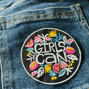 May include: A circular black embroidered patch featuring the white text GIRLS CAN in the center, surrounded by colorful floral motifs in shades of pink, yellow, and light blue, stitched onto a blue denim fabric background with visible seams.