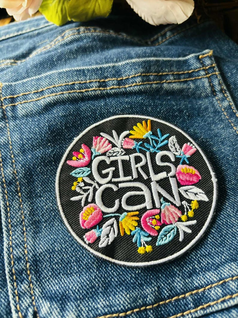 May include: A circular black embroidered patch featuring the white text GIRLS CAN in the center, surrounded by colorful floral motifs in shades of pink, yellow, and light blue, stitched onto a blue denim fabric background with visible seams.