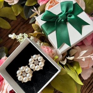 May include: A pair of pearl and rhinestone earrings in a white gift box with a green bow. The box is surrounded by pink and green flowers.