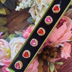 May include: A set of seven heart-shaped brooches in a green box. The brooches are gold-toned with red and pink rhinestones.