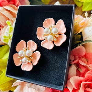 May include: A pair of light pink flower-shaped earrings with pearl and rhinestone accents, presented in a black box. The earrings have a gold-colored center and are set against a backdrop of pink and green flowers.
