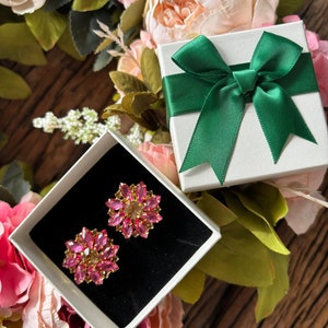 May include: A pair of pink flower-shaped earrings with gold accents are displayed in a white gift box with a green bow. The box is surrounded by pink flowers.