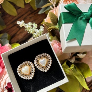 Könnte beinhalten: Eine weiße Geschenkbox mit einem grünen Satinband und Schleife. Die Box ist geöffnet und enthält zwei herzförmige Ohrringe mit Perlenbesatz.