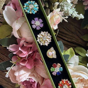 May include: A green box containing six jeweled flower brooches. The brooches feature various colors, including blue, purple, pink, gold, red, and green. The brooches are set against a black background and surrounded by a floral arrangement.