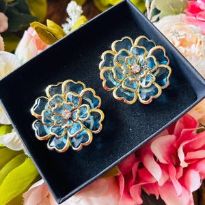 Op de afbeelding: Twee bloemvormige oorbellen met een goudkleurige rand en een lichtblauw middenstuk worden in een zwarte doos gepresenteerd. De oorbellen hebben een heldere, edelsteenachtige uitstraling en een kleine, heldere edelsteen in het midden. De achtergrond is gevuld met kleurrijke bloemen.