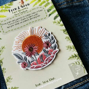 Puede incluir: Parche bordado con dise&ntilde;o de sol y flores en tonos naranja, rosa y blanco. Presentado en una tarjeta con texto: "Fern & Wren, Fast Upcycling, Slow Fashion". Dise&ntilde;ado para cubrir agujeros, manchas y desgarros.