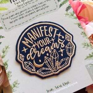 May include: A circular navy blue embroidered patch featuring gold thread text that reads "MANIFEST YOUR DREAMS" surrounded by stitched crystal, star, and crescent moon motifs. The patch is mounted on a branded card with fern illustrations.