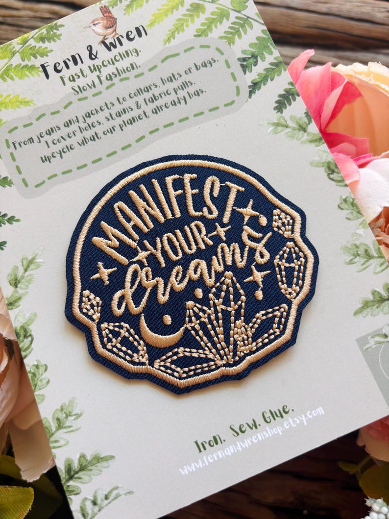 May include: A circular navy blue embroidered patch featuring gold thread text that reads "MANIFEST YOUR DREAMS" surrounded by stitched crystal, star, and crescent moon motifs. The patch is mounted on a branded card with fern illustrations.