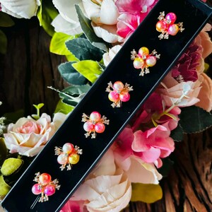 May include: A black box containing six floral-shaped hair pins. Each pin features a cluster of pink, yellow, and white pearls, accented with small, sparkling crystals. The pins are arranged against a backdrop of colorful flowers.