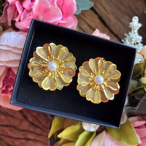 May include: A pair of gold-colored flower-shaped earrings with a pearl-like center, presented in a black box. The earrings have a delicate, mesh-like design. The background features pink flowers and greenery.