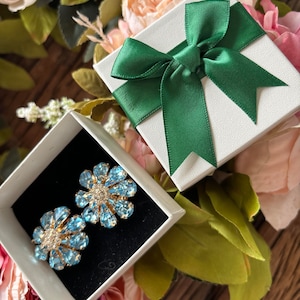 May include: A pair of blue flower-shaped earrings with clear rhinestones, displayed in a white gift box with a green satin ribbon.
