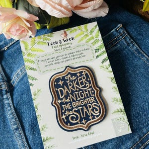 May include: A navy blue embroidered patch with a decorative border and gold lettering that reads, The DARKER the NIGHT the BRIGHTER the STARS. The patch is mounted on a white card featuring green fern illustrations and text about upcycling.